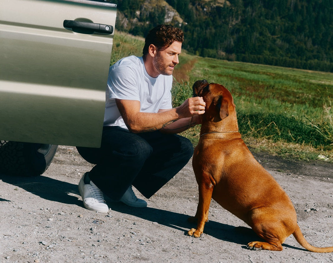 A man crouching beside a parked vehicle outdoors, interacting with a brown dog in a scenic natural setting.