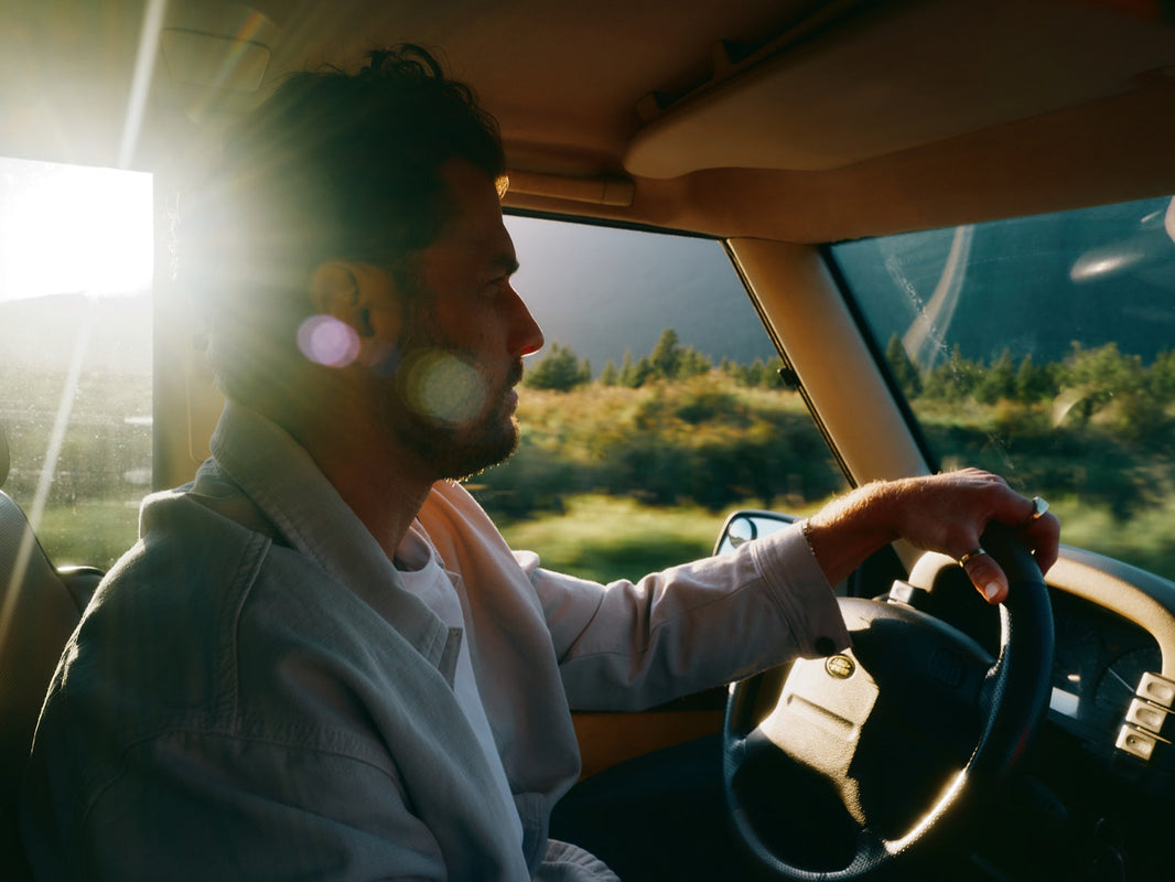 A man driving a car with sunlight streaming through the window, highlighting a scenic road and lush greenery outside.
