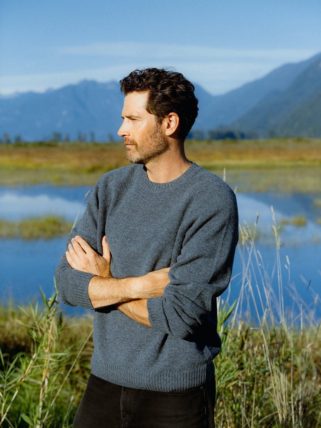 A man standing outdoors near a lake and mountains, reflecting thoughtfully with arms crossed.