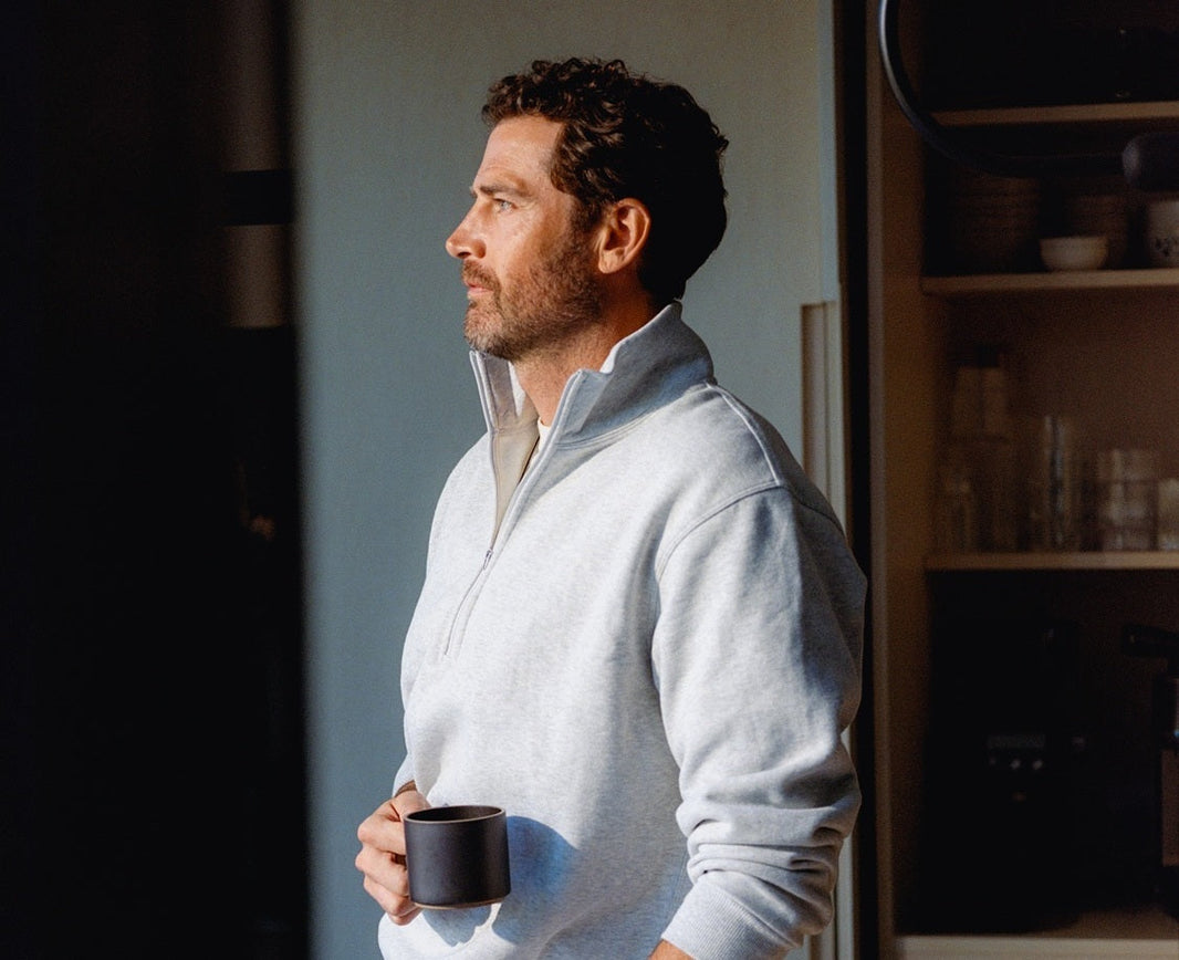 A man in a light sweater holding a coffee cup, standing thoughtfully near a kitchen counter.